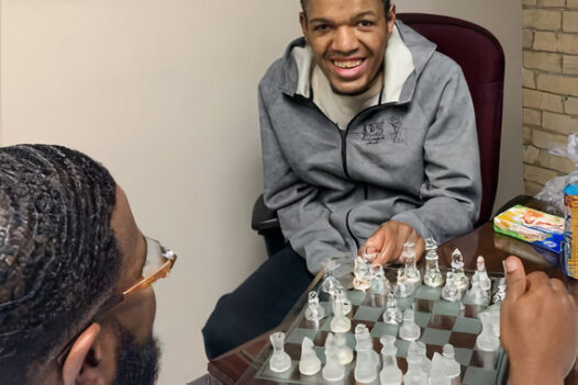 A young man with a disability plays chess and smiles at the camera.
