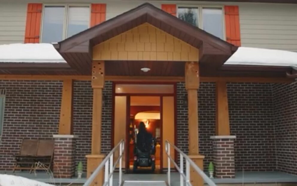 Person in wheelchair entering a home, in foreground is the ramp to the front door.