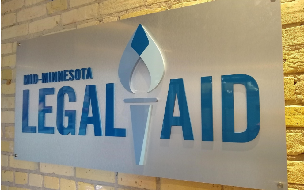 Lobby sign on shiny silver background reads Mid-Minnesota Legal Aid in blue raised lettering with torch symbol.