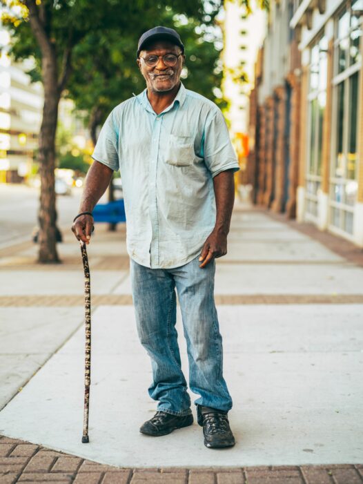 An older man wearing a baseball cap, short-sleeved shirt and jeans stands on a sidewalk with a cane.