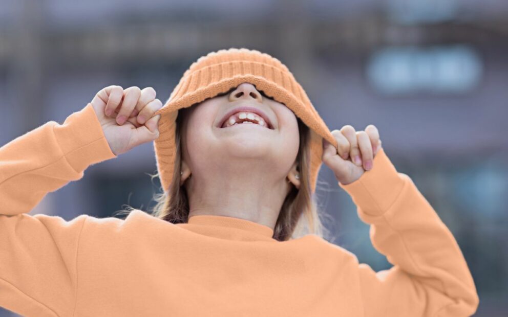 Eight-year-old girl in peach sweater and matching peach-colored cap, smiles, pulling cap over her eyes.