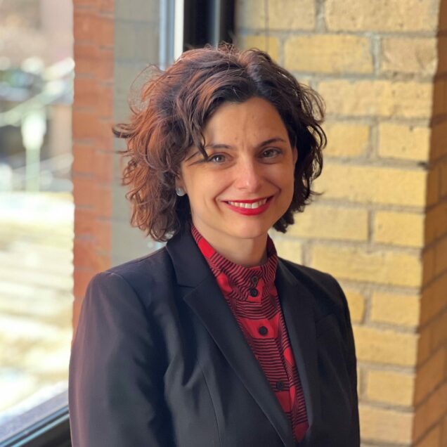 Abigail, wearing a red blouse and black blazer, stands in front of a window and smiles at the camera.