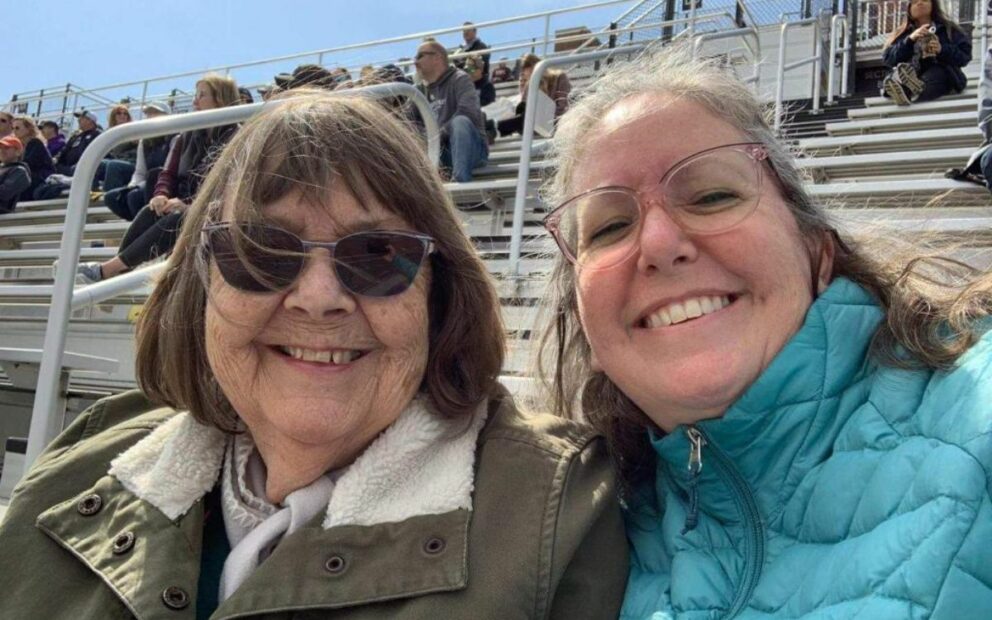 Wearing jackets, Grandmother and adult daughter smile in stands at outdoor track meet.