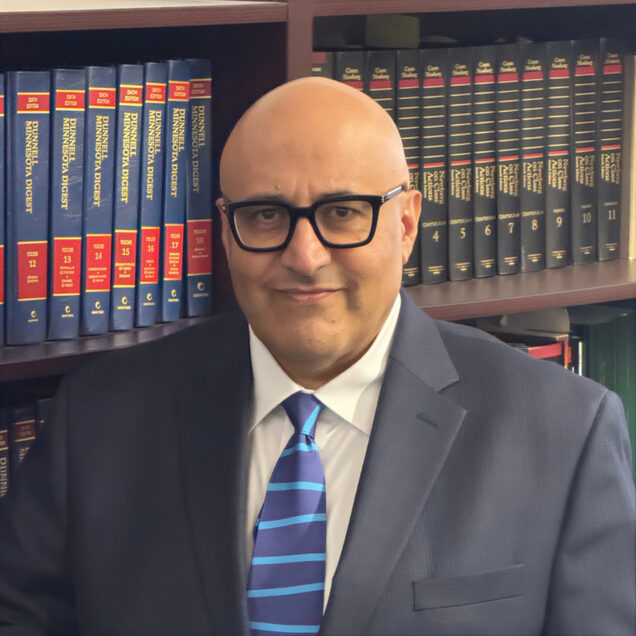 Professional headshot of a man in front of book case, dressed in dark suit, white shirt, maroon tie.