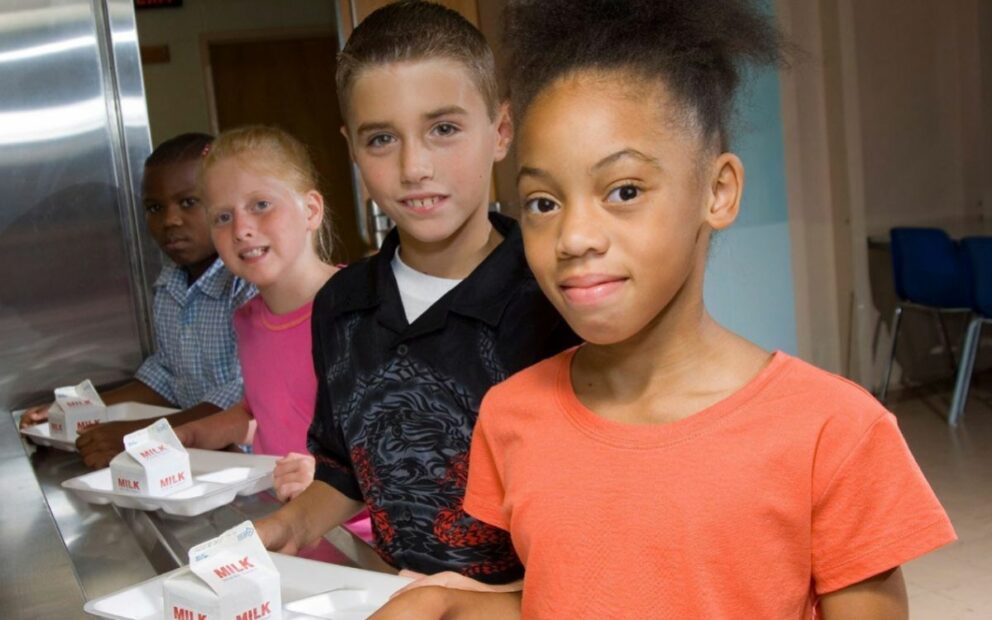 Four elementary-aged students in lunch line smile at the camera.