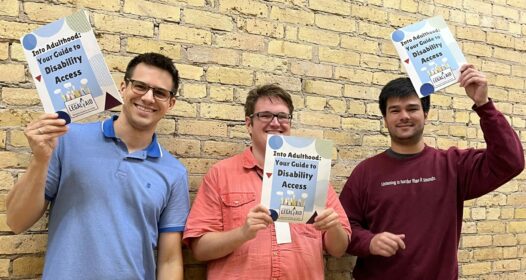 Three Legal Aid staff members stand against a brick wall and hold up copies of the Into Adulthood Guide.