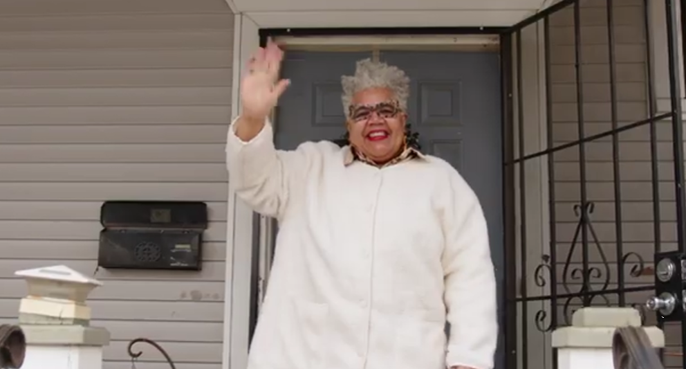 Woman stands on porch and waves "hello" toward the street.