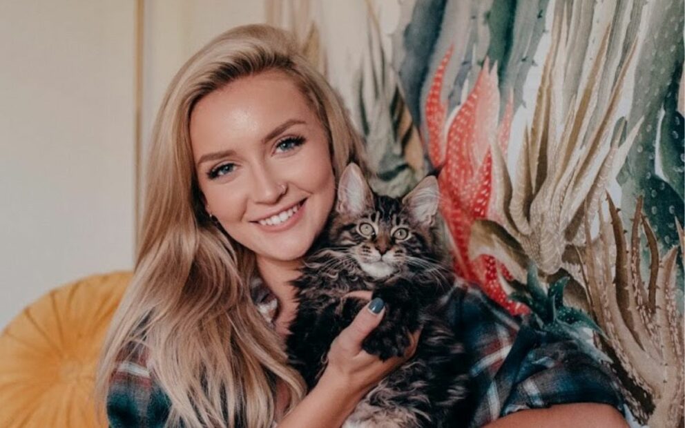 Young, blonde woman smiles while holding cat with dark grey fur close to her face.