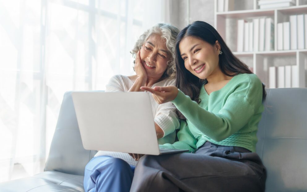 An older woman and her adult daughter sit together on a couch, looking at a laptop screen.