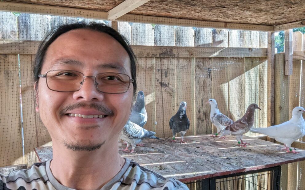 Man wearing glasses and a smile stands in bird coop with pigeons on elevated platform in background.