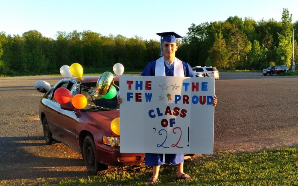 Student in cap and gown stands in front of car decorated with balloons, holding sign, "The few the proud Class of 2022."