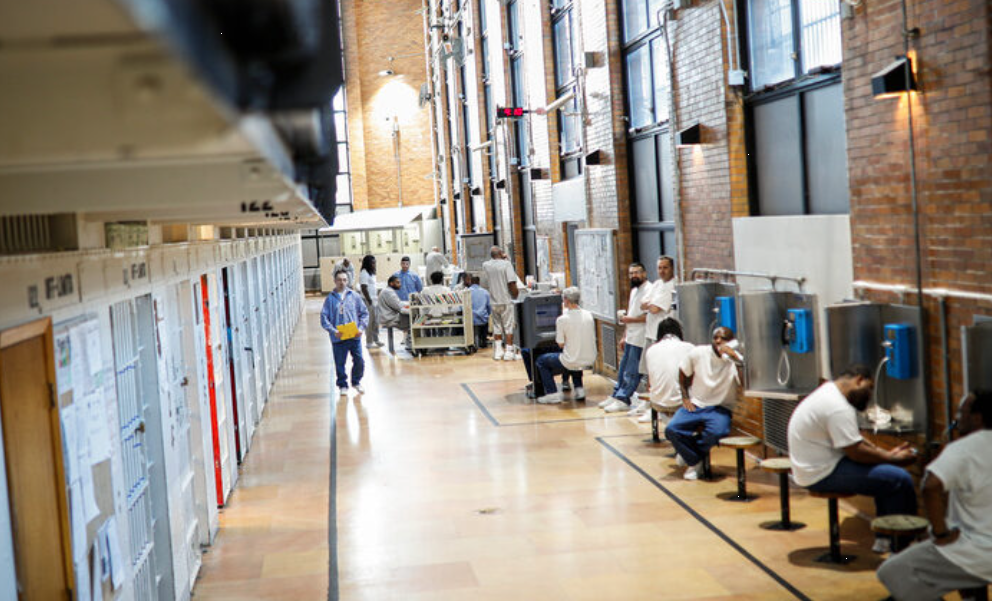 Inmates sit at phone stations on right side of hallway. Some are in the hallway, lined by cells on left side.