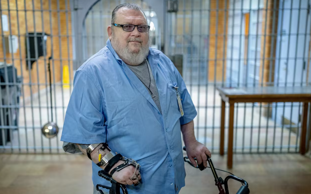 Man with glasses, greyed beard, wearing blue, long-sleeved shirt, stands in front of prison cell.