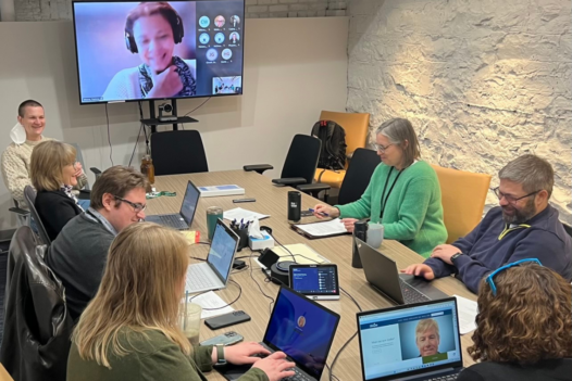 Staff members seated around a large conference table. Remote attendees appear on large screen in background.