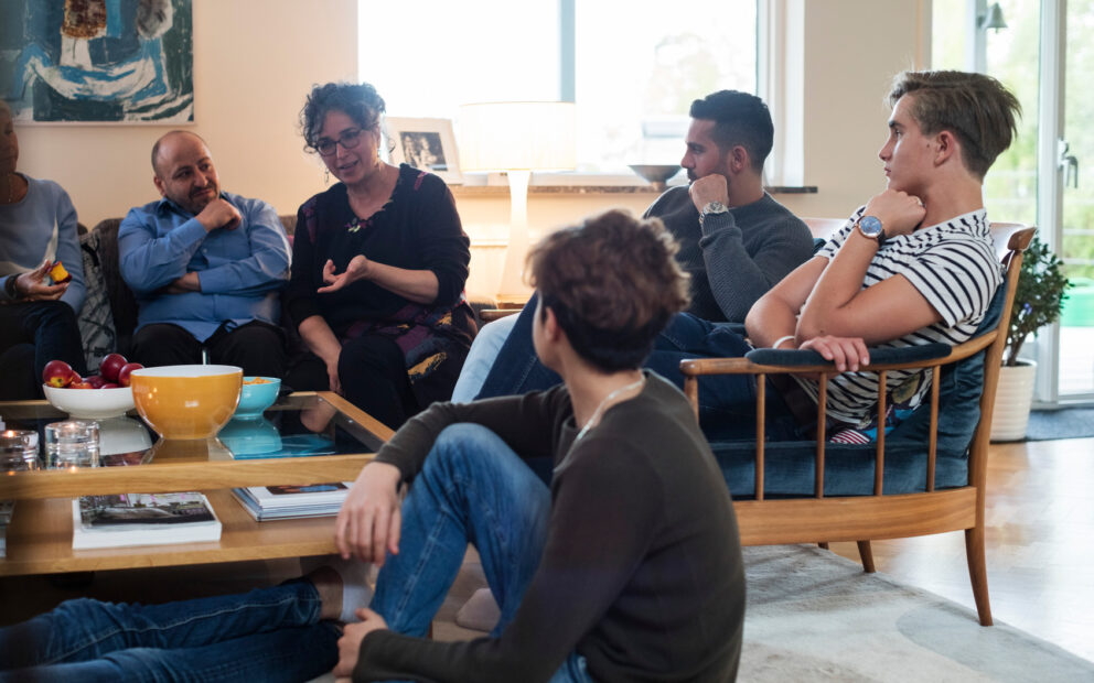 Multigenerational family and friends sit on sofa, chairs and on the floor conversing living room