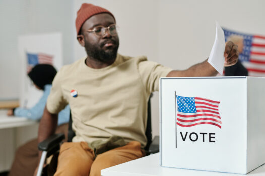 Box for ballots in foreground. In the background a man in a wheelchair extends arm to place ballot into ballot box.