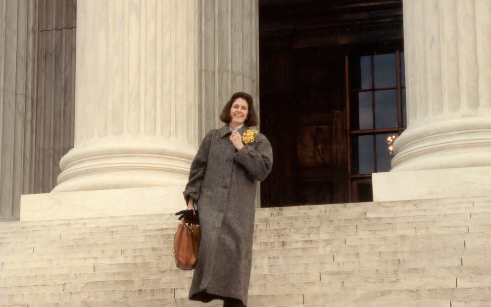 Woman with chin-length brown hair in long tweed coat with stands smiling on steps of the U.S. Supreme Court