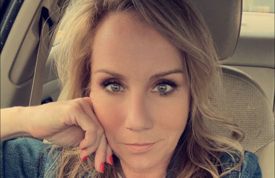 Woman with dark blonde shoulder length hair sits in a car, hand to her cheek, elbow resting on window fram