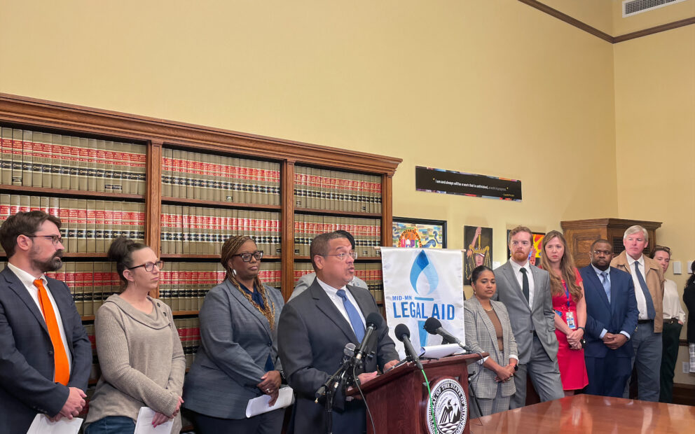 Lawyers stand behind man at podium with microphones and banner of Mid-Minnesota Legal Aid