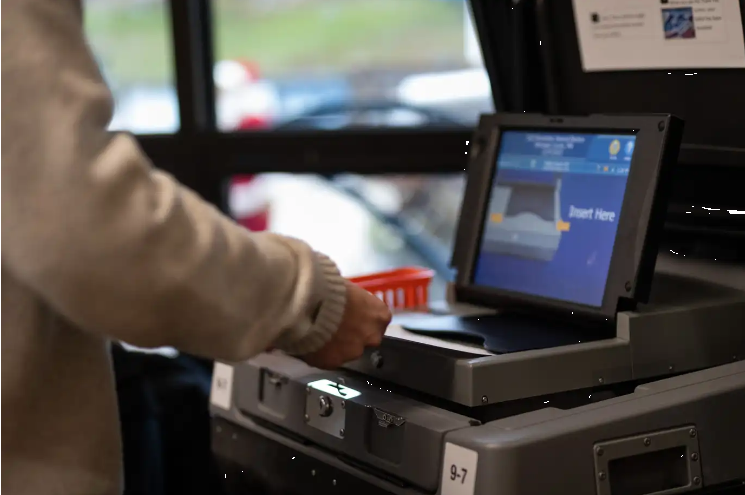 Person in a beige coat puts a ballot into a voting machine which has a digital screen on top