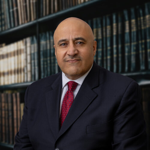 Professional headshot of a man in front of book case, dressed in dark suit, white shirt, maroon tie.