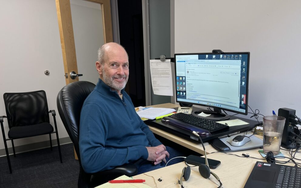 A man in a blue sweater turns to camera smiling, while seated at his office desk, computer screen in background and legal notepads and headset scattered on his side desk