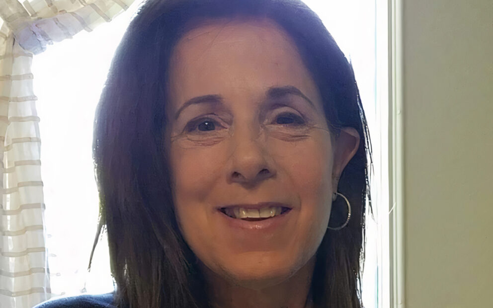 Woman with dark brown, shoulder-length hair smiles. Sun shines in from the window behind her.