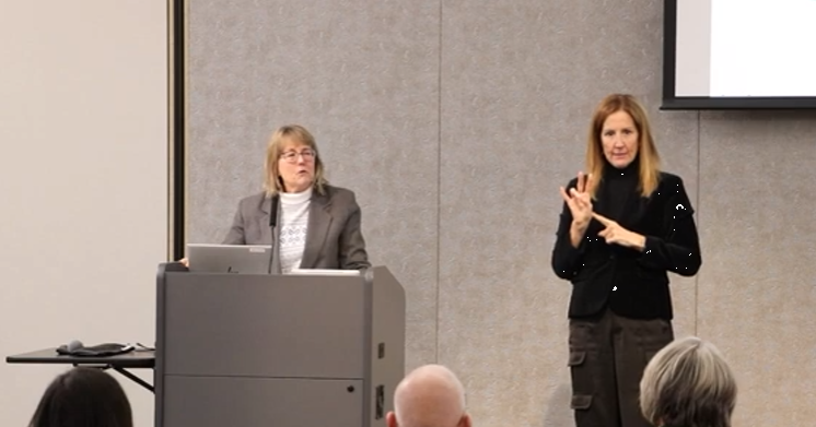 Woman in white shirt and tan blazer at lectern speaks while a woman in black sweater and black pants stands next to her uses American Sign Language to interpret her remarks