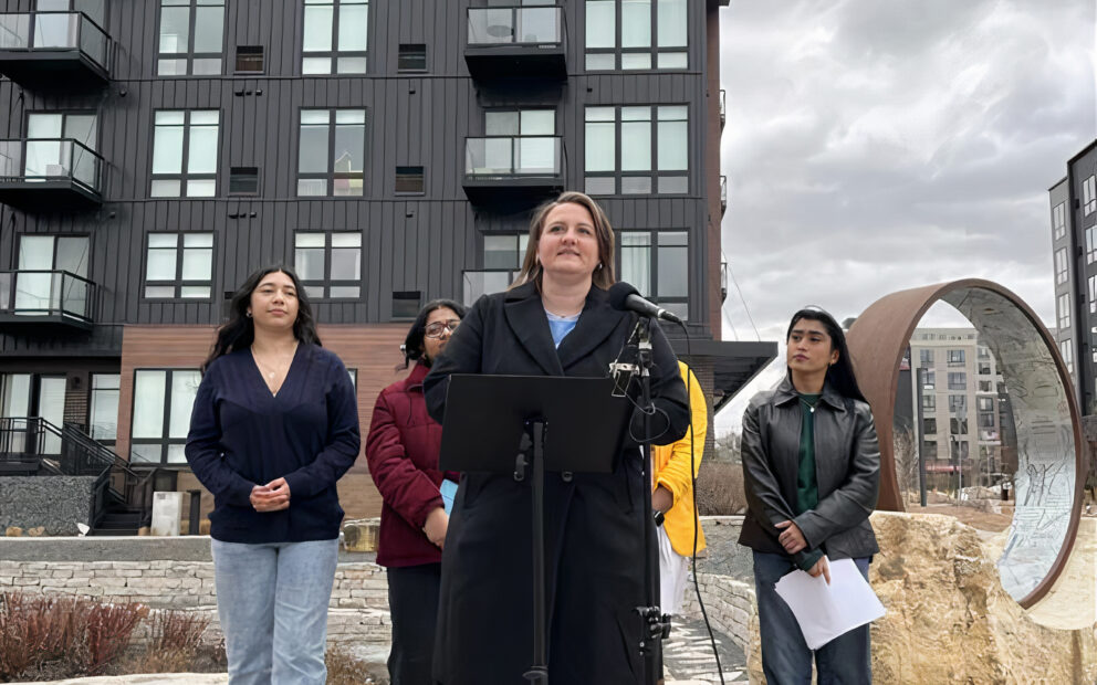 Outdoor news conference scene with a woman at microphone; behind her are four other women standing behind her