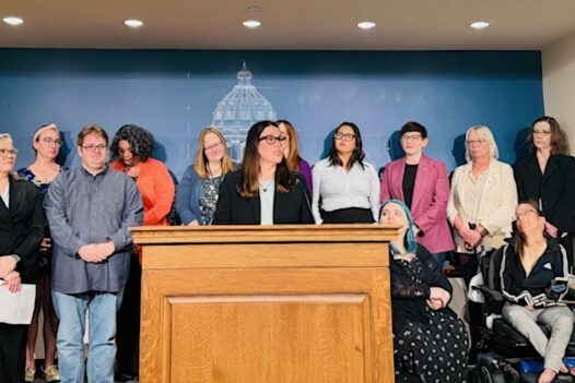 News conference setting with outline of Capitol on wall in background. A woman is speaking at a microphone with others standing behind her.
