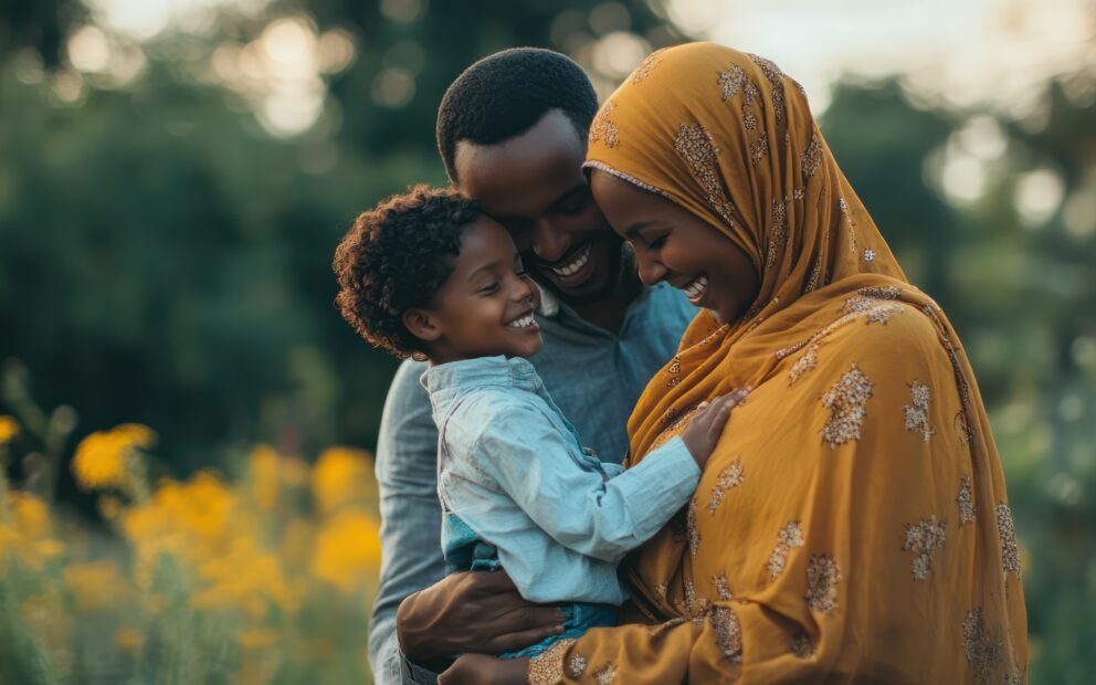 Father and mother face their toddler-age child as they hold him. All are smiling. Mother is wearing a yellow hijab. A field of yellow flowers in the background on a sunny day.