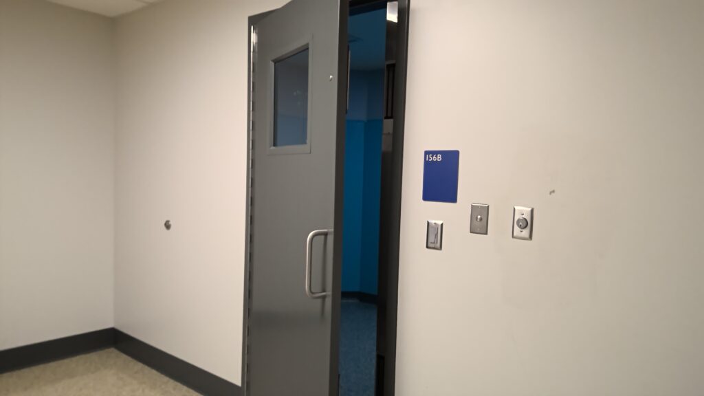 Metal door with observation window towards the top. A blue sign on the adjacent wall reads 156B
