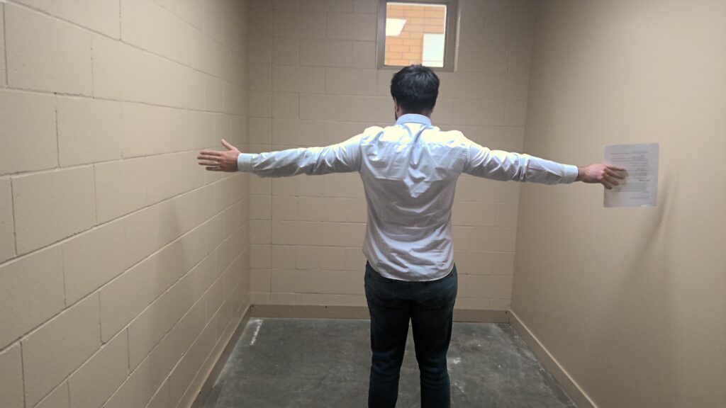 A small room with beige cinder block walls and concrete floor. An adult stands in the center of the room with arms stretched out to the side to show scale. The room is only slightly wider than the adult's arm span.
