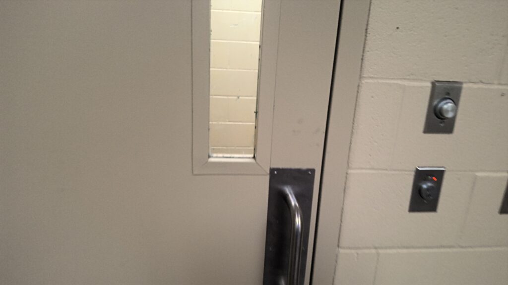 Photograph of a closed beige door with a narrow vertical window and a metal handle, set in a cinder block wall. Adjacent wall features two metal switches.