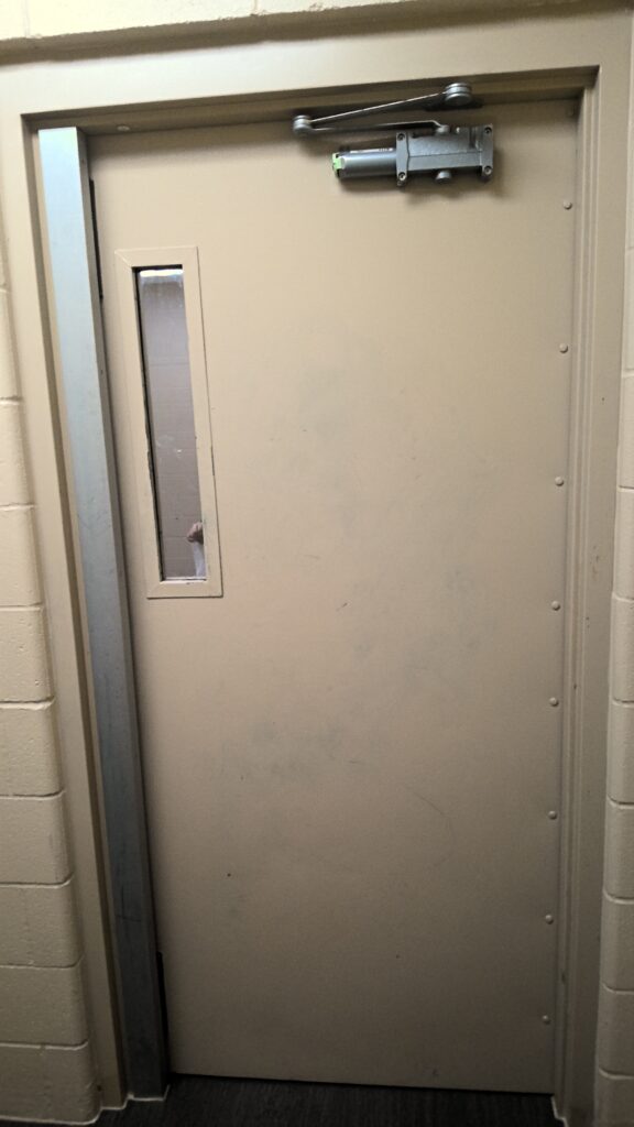 A beige metal door with a door closer mechanism and a narrow observation window.