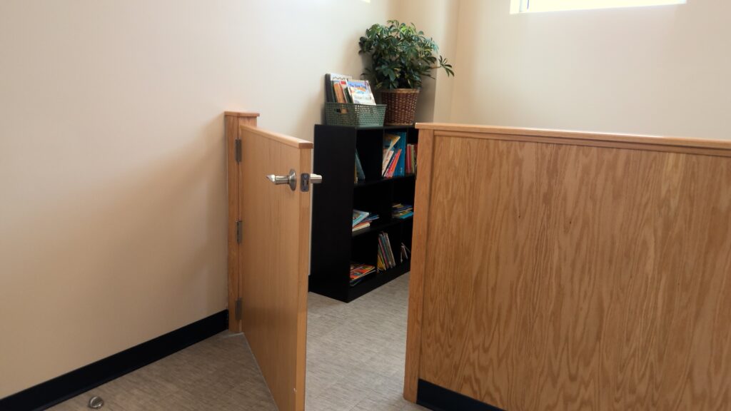 A room with a wood half-wall barrier with an open door. On the other side of the barrier is a black bookshelf with children's books and a house plant.