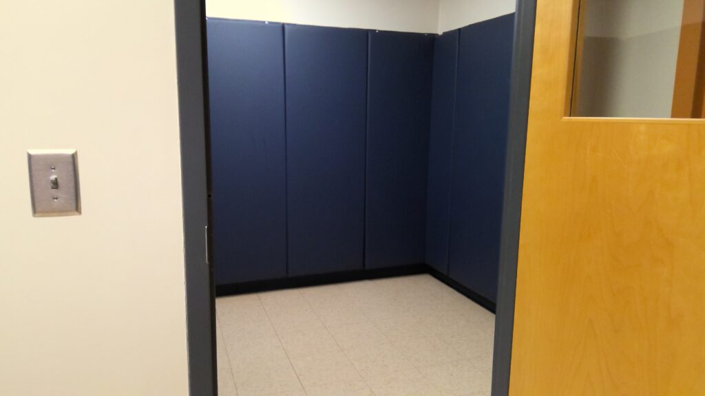 Viewed through a doorway, a small room with padded blue walls and a white un-carpeted floor. The wooden door has an observation window at the top and is wide open.