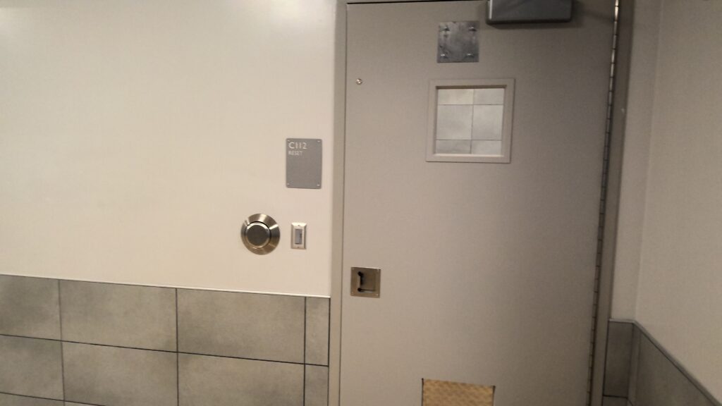 Photograph of a closed, solid gray door with a small square window covered by a metal plate, set in a tiled hallway with light gray walls. A round metal fixture and a sign labeled "C112" are mounted on the wall next to the door, indicating a secured or restricted area.