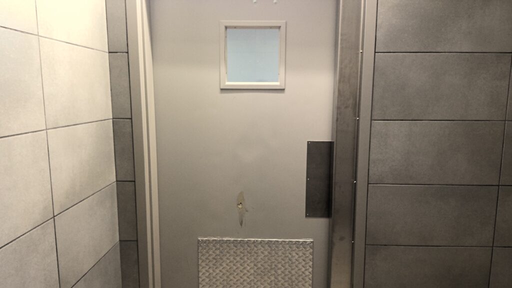 A room with slate gray tiled walls and a metal door with a metal kick plate and small observation window.