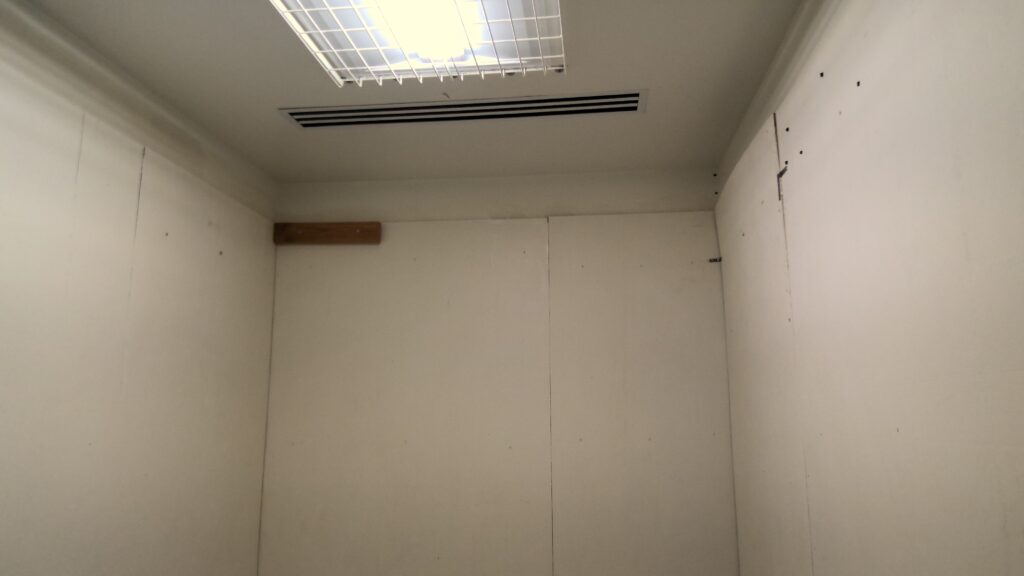 Room with white paneling, grated light fixture, and ceiling vent.