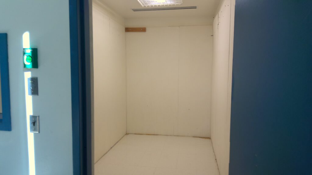Room with white paneling and grated ceiling light, viewed through open doorway.