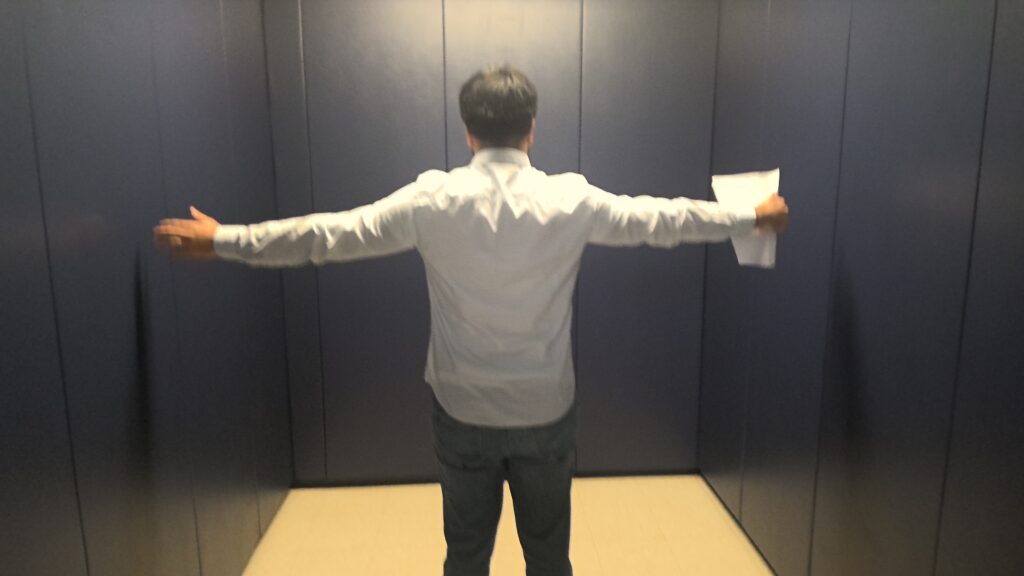 An adult stands in the middle of a narrow room with blue padded walls. Their arms are extended out to their side to show scale. Their arm span covers almost the full width of the room with only a few inches of space between their fingertips and the walls.