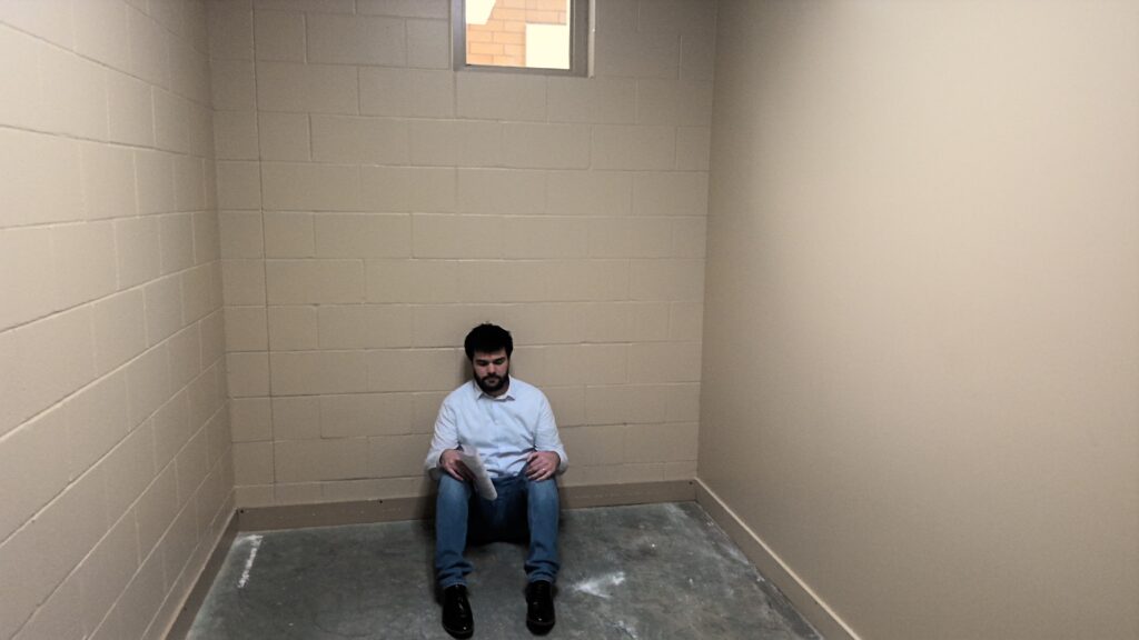 An adult sits on the floor in a narrow room with two cinder block walls and one plaster wall. The floor is unfinished concrete with white staining. There is a small window towards the top of the wall.