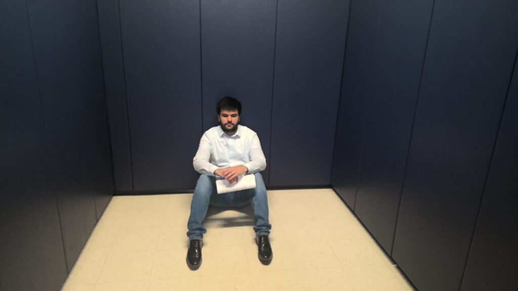 An adult sitting on the floor with their back against the wall in a room with blue padded walls.