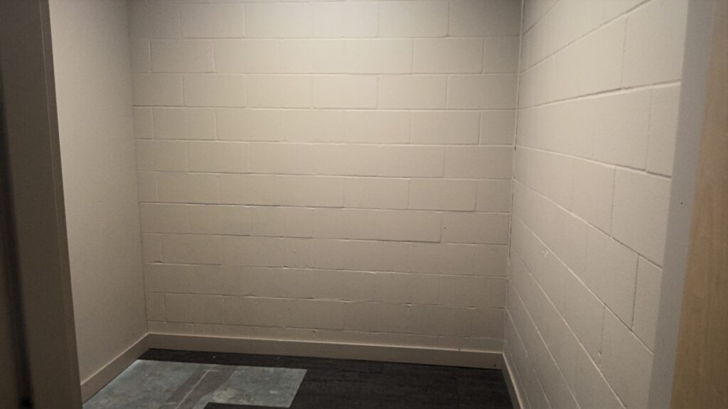 A small room with beige cinder block walls. Squares of thin commercial carpet cover only part of the floor, leaving uncovered sections of cement.