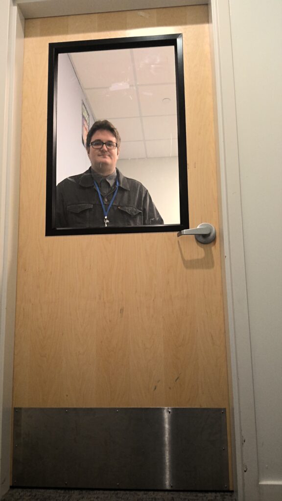 A wooden door with metal kick panel along the bottom and a large window in the upper half. An adult male looks at the camera from an observation window. His upper half of the body is visible to the viewer.