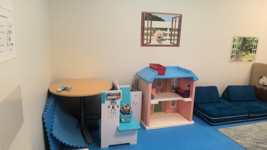 A room with a pink dollhouse, thick floor cushions, and a small table. Visuals on the wall appear to create windows to farm animals outside the room.