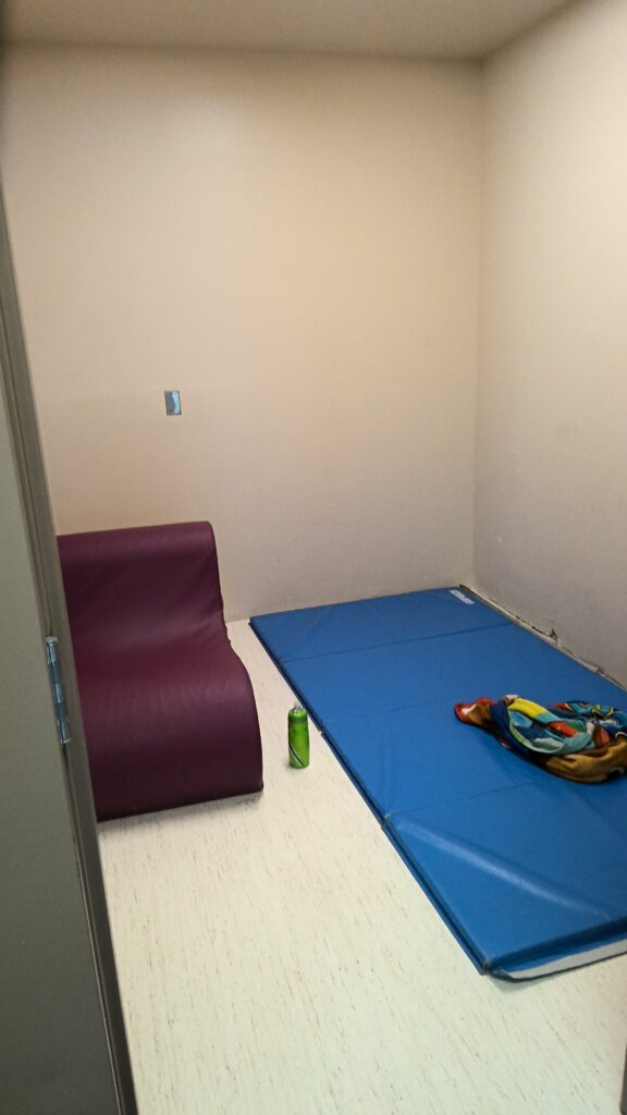 Photograph of a small, sparsely furnished room with a blue mat and colorful blanket placed on the floor in one corner. A purple cushioned seat and a green water bottle are positioned near the mat, with plain beige walls and light-colored flooring.