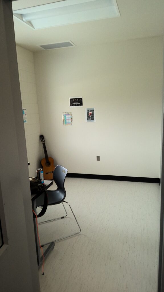 Room with beige walls and light flooring. A portable desk and chair, guitar, and small posters on the wall.
