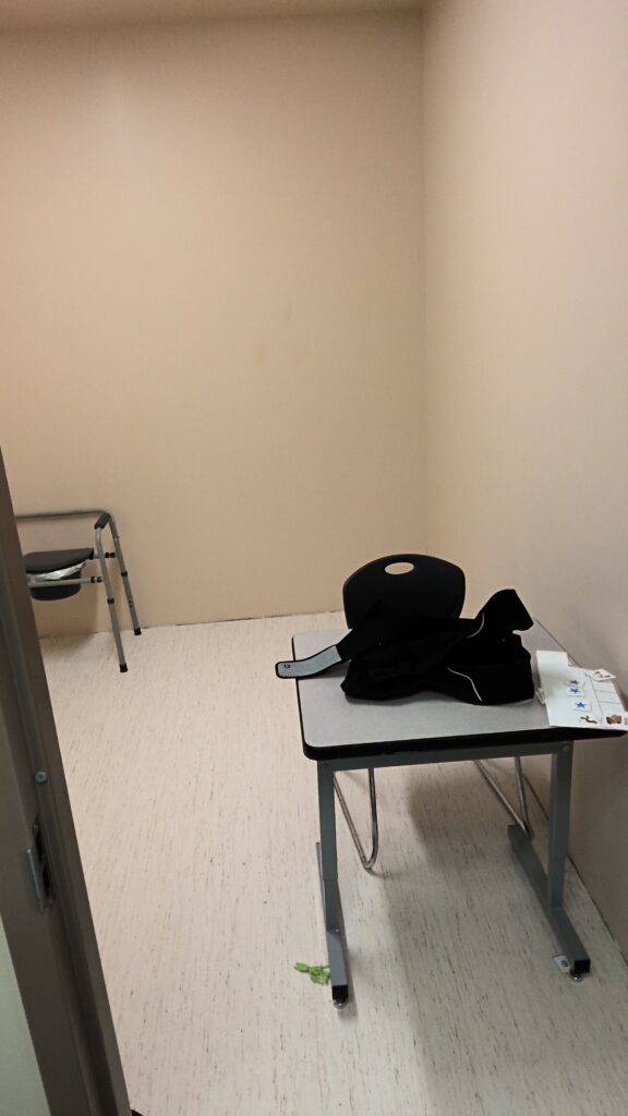 Room with beige walls and white flooring, desk and chair, and a portable toilet.
